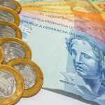 Imagem com moedas e cédulas de reais espalhadas, incluindo notas de 2, 5, 10, 20, 50 e 100 reais, representando o pagamento de RPVs pelo CJF.