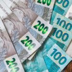 Notas de 200 e 100 reais espalhadas sobre a mesa ao lado de moedas brasileiras, representando dinheiro, pagamento e recebimento de valores como RPV e precatórios.