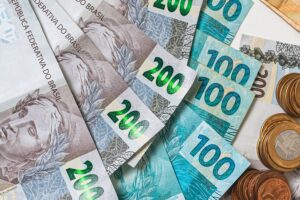 Notas de 200 e 100 reais espalhadas sobre a mesa ao lado de moedas brasileiras, representando dinheiro, pagamento e recebimento de valores como RPV e precatórios.