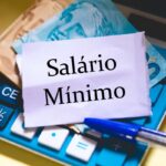 Nota com a frase ‘Salário Mínimo’ sobre calculadora, caneta e cédulas de real, representando o valor do piso nacional